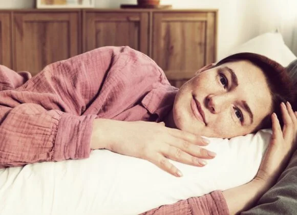 The Best Sleep Positions to Alleviate Neck Pain: Side Sleeping Tips A person with a short haircut rests on a pillow, lying on a bed in a softly lit room and looking at the camera, showcasing proper spinal alignment during sleep.
