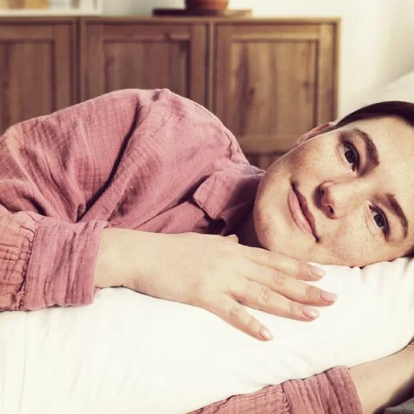 A person with a short haircut rests on a pillow, lying on a bed in a softly lit room and looking at the camera, showcasing proper spinal alignment during sleep.