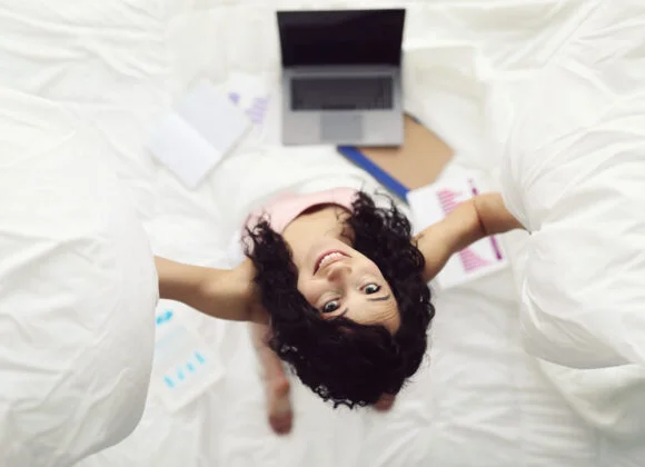A person in a light pink top looks up while holding pillows. A laptop, notepads, and papers with graphs are spread around on the bed.