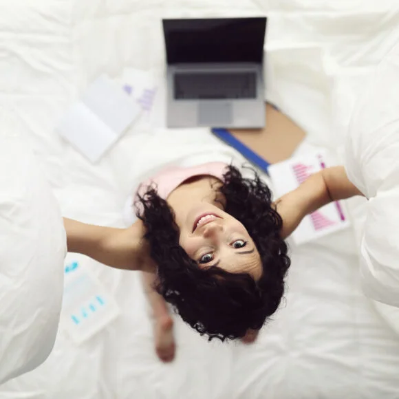 A person in a light pink top looks up while holding pillows. A laptop, notepads, and papers with graphs are spread around on the bed.