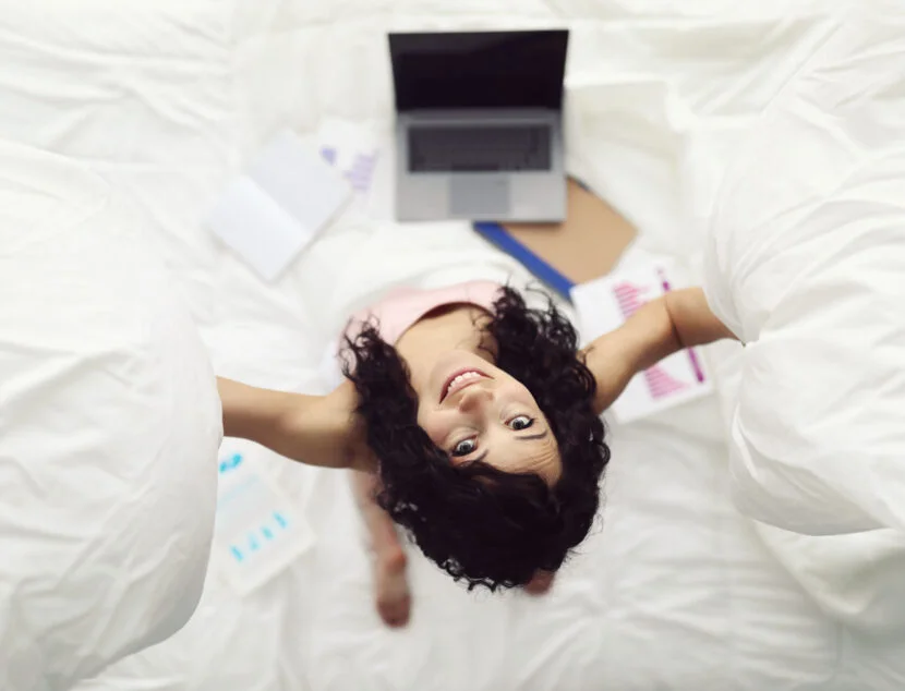 A person in a light pink top looks up while holding pillows. A laptop, notepads, and papers with graphs are spread around on the bed.