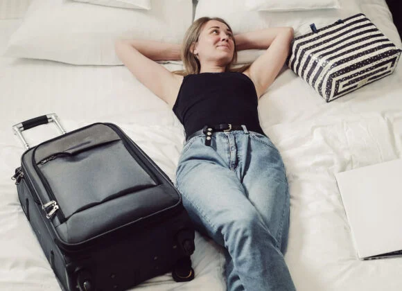 A person in a black tank top and jeans is lying on a bed beside a suitcase, a striped bag, and a closed laptop.