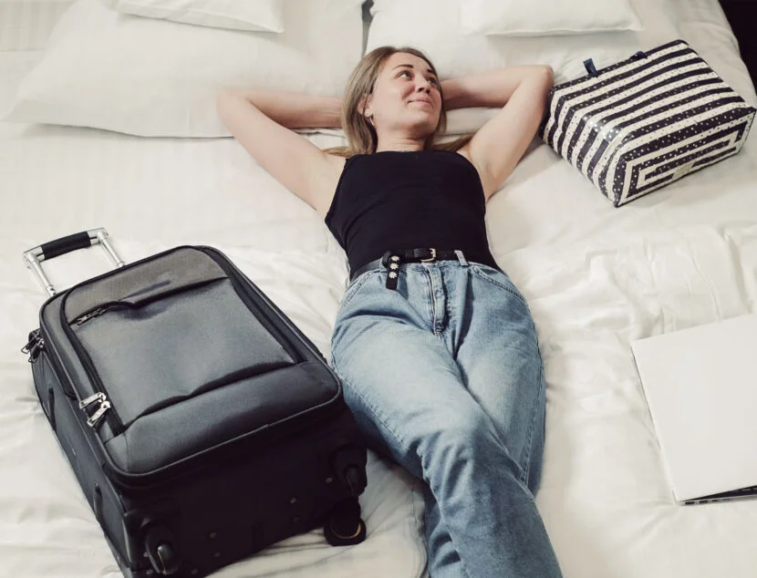 A person in a black tank top and jeans is lying on a bed beside a suitcase, a striped bag, and a closed laptop.
