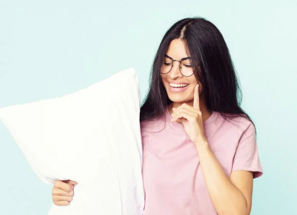 A woman with long dark hair and glasses smiles while holding a white pillow and touching her face. She is wearing a pink T-shirt, and the background is light blue.