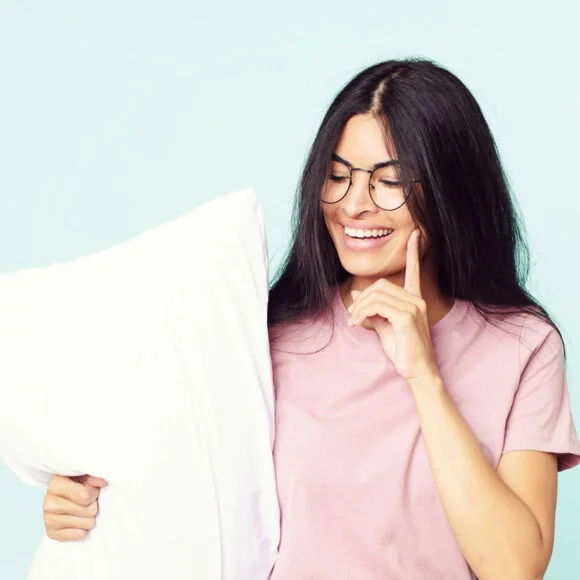 A woman with long dark hair and glasses smiles while holding a white pillow and touching her face. She is wearing a pink T-shirt, and the background is light blue.