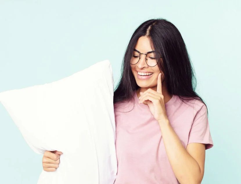 A woman with long dark hair and glasses smiles while holding a white pillow and touching her face. She is wearing a pink T-shirt, and the background is light blue.