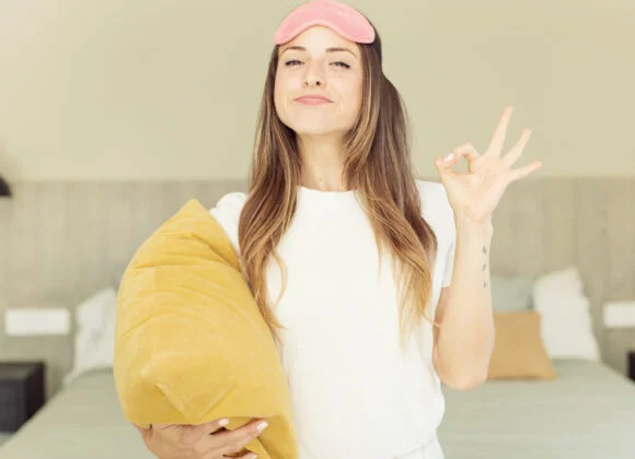A person wearing a white shirt and a pink sleep mask stands in a bedroom holding a yellow pillow, gesturing an "OK" sign with their hand.