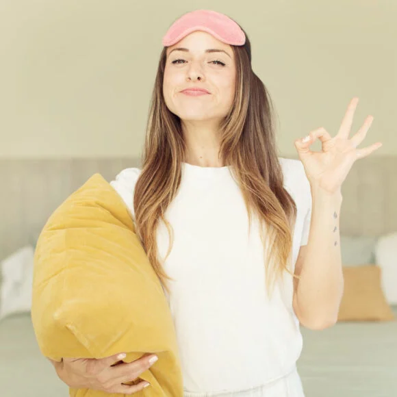 A person wearing a white shirt and a pink sleep mask stands in a bedroom holding a yellow pillow, gesturing an "OK" sign with their hand.