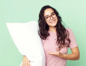 A person with long curly hair and glasses holds a white pillow and gestures towards it while smiling. They are wearing a pink shirt and standing against a light green background.