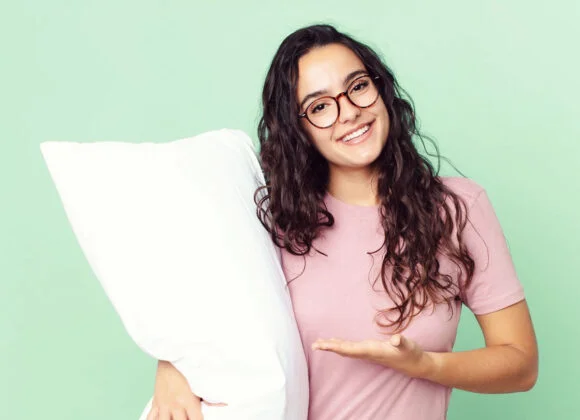 Cervical Pillows Explained: The Key to Pain-Free Sleep A person with long curly hair and glasses holds a white pillow and gestures towards it while smiling. They are wearing a pink shirt and standing against a light green background.