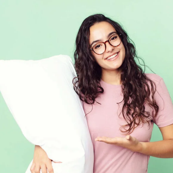 A person with long curly hair and glasses holds a white pillow and gestures towards it while smiling. They are wearing a pink shirt and standing against a light green background.