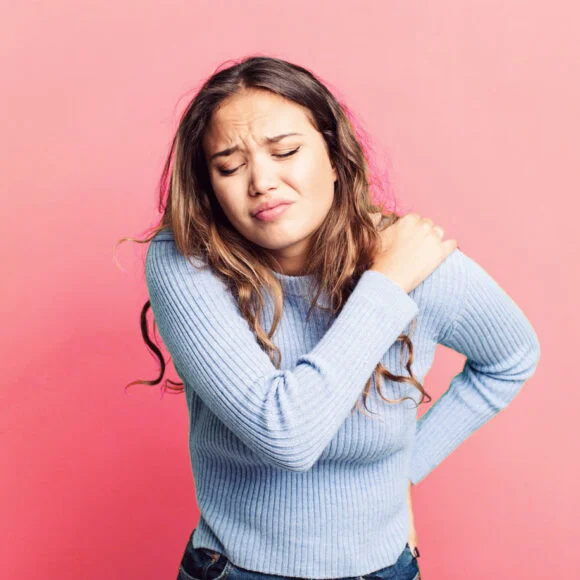 A person with long hair in a light blue sweater holds their left shoulder with their right hand while showing a pained expression against a pink background.