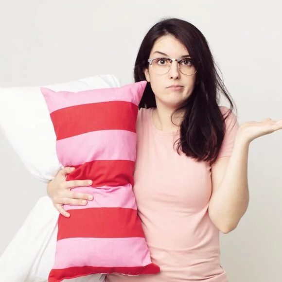 A woman with glasses shrugs while holding a striped pillow and a white pillow against a plain background.
