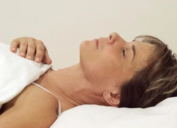 The Benefits of Using Contoured Pillows for Neck Pain Relief A person lies in bed on their back with eyes closed and one hand on the blanket. The background is plain.