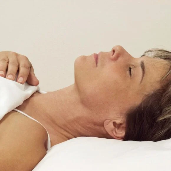 A person lies in bed on their back with eyes closed and one hand on the blanket. The background is plain.