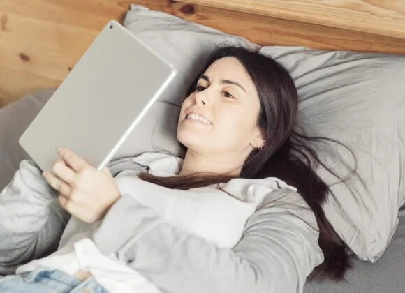 A woman lies on a bed, propped up by pillows, while using a tablet.