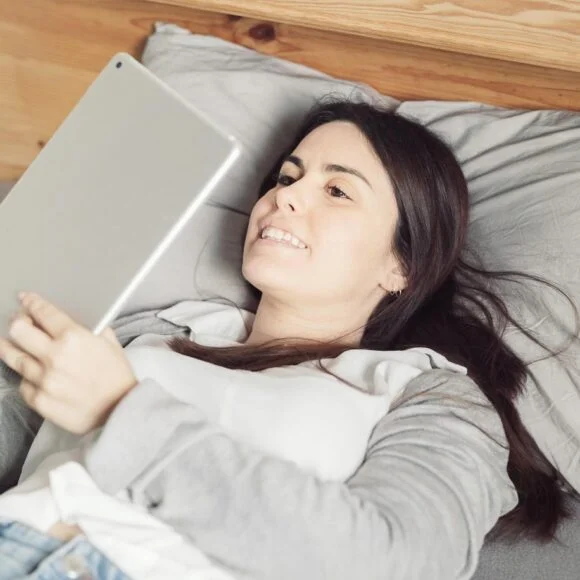 A woman lies on a bed, propped up by pillows, while using a tablet.