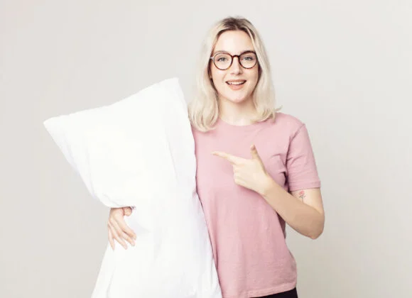 Person with glasses and a pink shirt smiling, holding a large white pillow with one hand and pointing at it with the other.
