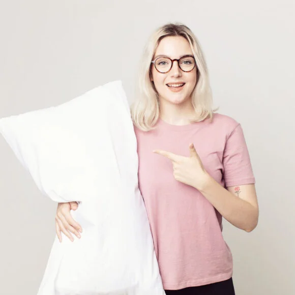 Person with glasses and a pink shirt smiling, holding a large white pillow with one hand and pointing at it with the other.