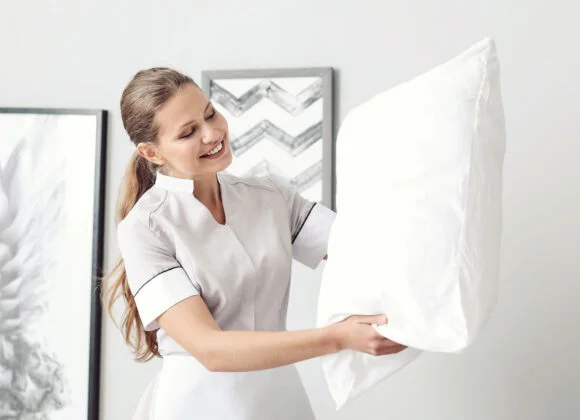 A smiling woman in a grey and white uniform holds a white pillow in a modern, brightly lit room with framed artwork on the wall.