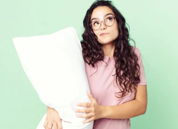 Woman with long curly hair and glasses holds a large white pillow, standing against a light green background. She is wearing a pink shirt and looking slightly upwards.