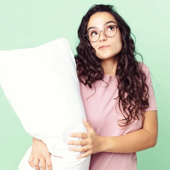 Woman with long curly hair and glasses holds a large white pillow, standing against a light green background. She is wearing a pink shirt and looking slightly upwards.