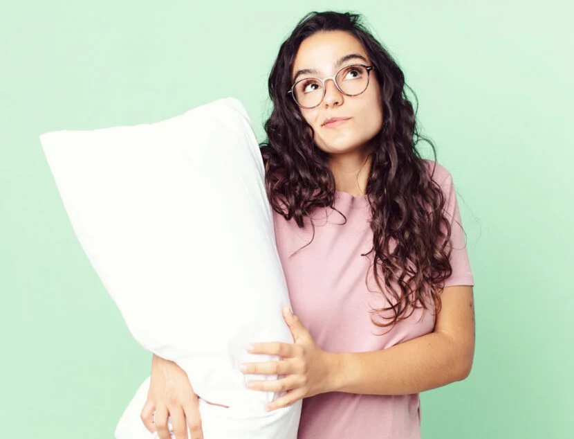 Woman with long curly hair and glasses holds a large white pillow, standing against a light green background. She is wearing a pink shirt and looking slightly upwards.