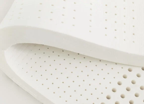 Close-up of a white, perforated foam mattress topper with small round holes uniformly distributed across its surface. The material appears flexible and slightly folded at the corner.