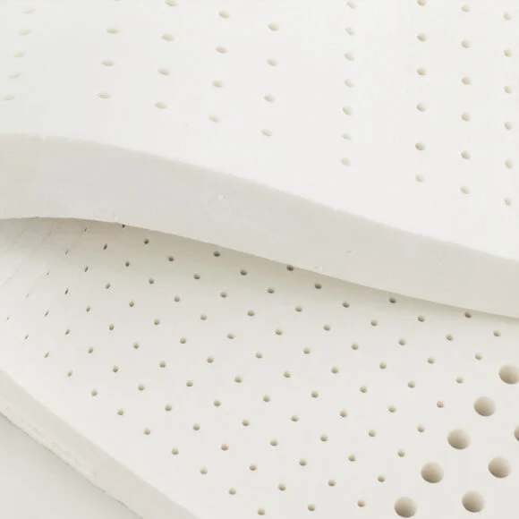 Close-up of a white, perforated foam mattress topper with small round holes uniformly distributed across its surface. The material appears flexible and slightly folded at the corner.
