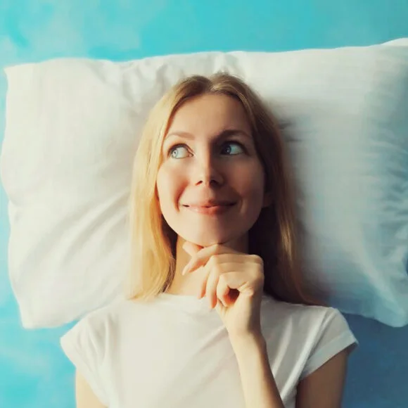 A woman with long blond hair lies on a white pillow, looking off to the side with a thoughtful expression. She is wearing a white shirt and the background is light blue.