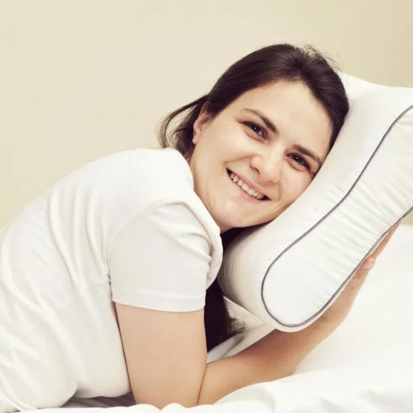 A person in a white shirt is lying on a bed, smiling, with their head resting on a white pillow to prevent neck pain from sleeping