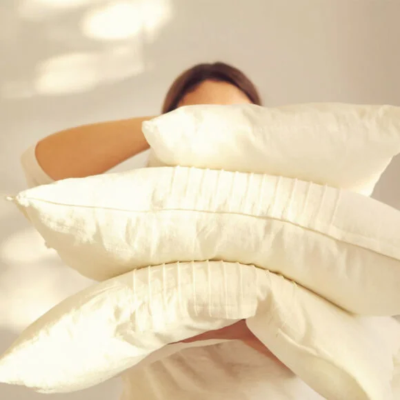 Person holding four off-white pillows in front of their face with a neutral background.