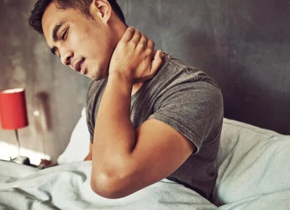 A man sits in bed, holding his neck with his right hand, appearing to experience neck pain. A red lamp is visible in the background.