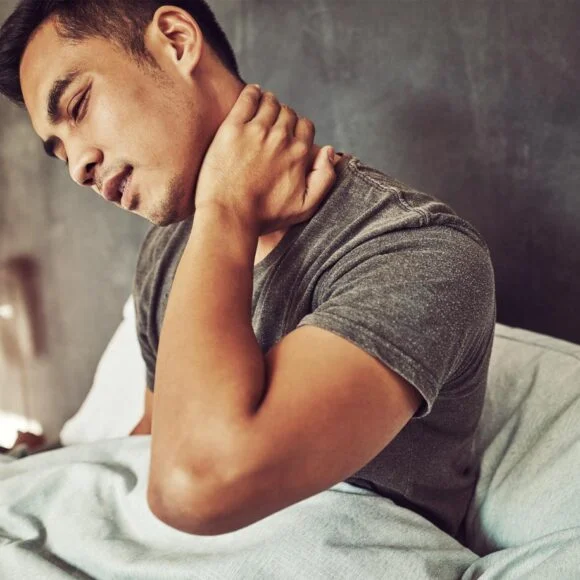 A man sits in bed, holding his neck with his right hand, appearing to experience neck pain. A red lamp is visible in the background.
