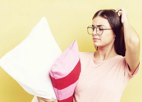 A woman with glasses and a pink shirt holds two pillows, one white and one pink with red stripes, while scratching her head in a puzzled manner. Yellow background.
