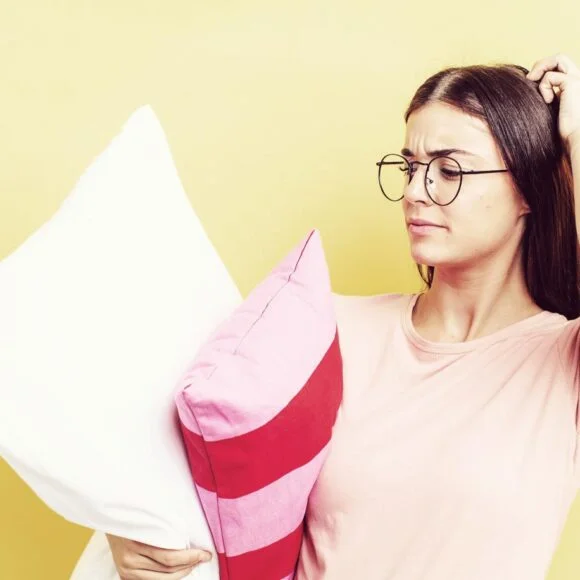 A woman with glasses and a pink shirt holds two pillows, one white and one pink with red stripes, while scratching her head in a puzzled manner. Yellow background.
