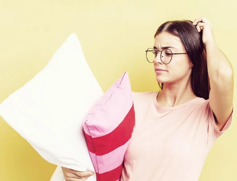 Orthopedic Pillows: What Are They and Do You Need One? A woman with glasses and a pink shirt holds two pillows, one white and one pink with red stripes, while scratching her head in a puzzled manner. Yellow background.
