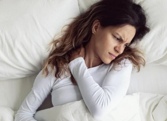How Pillow Size Affects Neck Pain: Choosing the Right Fit A woman lying in bed with a pained expression, holding her neck with one hand, appears to be experiencing discomfort or stiffness. She has long hair and is dressed in a white long-sleeve shirt.