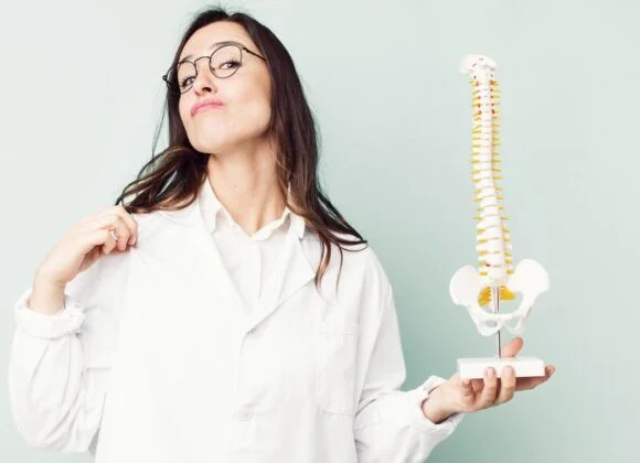 A woman in a lab coat points to her shoulder while holding a spine model.