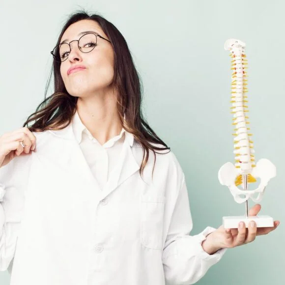 A woman in a lab coat points to her shoulder while holding a spine model.