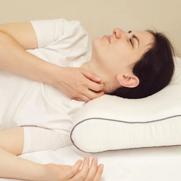 A person lying on a bed with a white pillow, holding their neck and wincing in pain due to improper pillow thickness.
