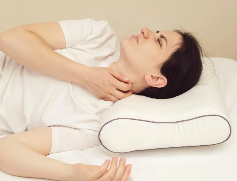 A person lying on a bed with a white pillow, holding their neck and wincing in pain due to improper pillow thickness.