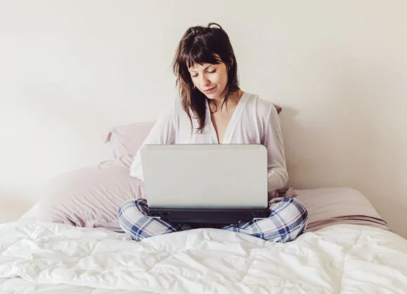A person sits cross-legged on a bed, using a laptop. The background is plain with a pillow visible.