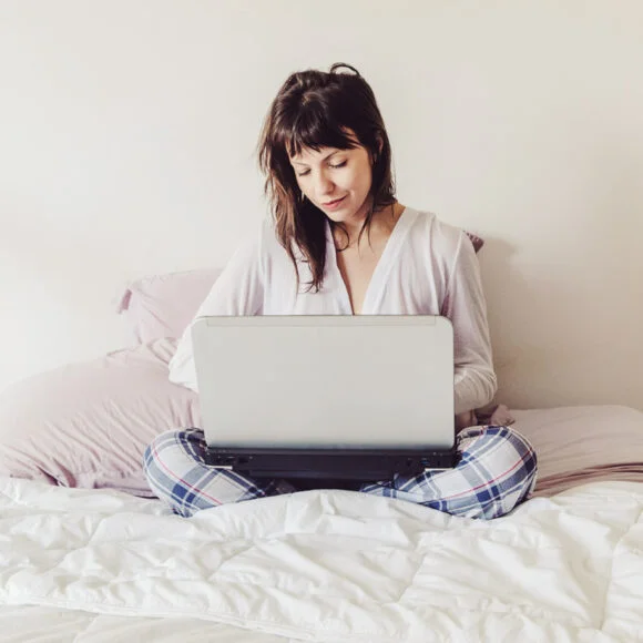A person sits cross-legged on a bed, using a laptop. The background is plain with a pillow visible.