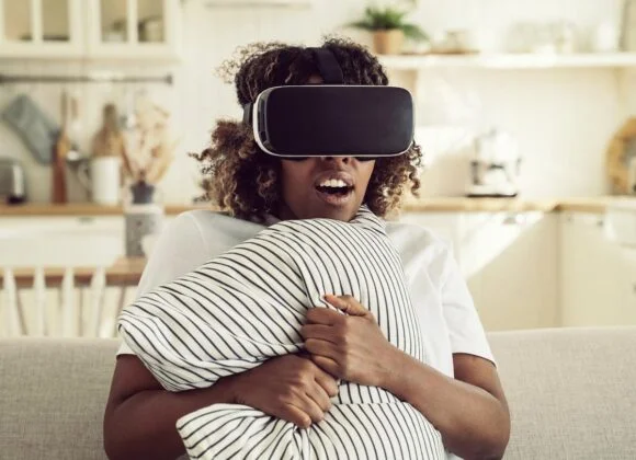 A person sitting on a couch wearing a virtual reality headset, holding a striped pillow while experiencing VR content in a cozy, well-lit living room.