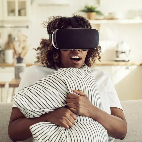 A person sitting on a couch wearing a virtual reality headset, holding a striped pillow while experiencing VR content in a cozy, well-lit living room.