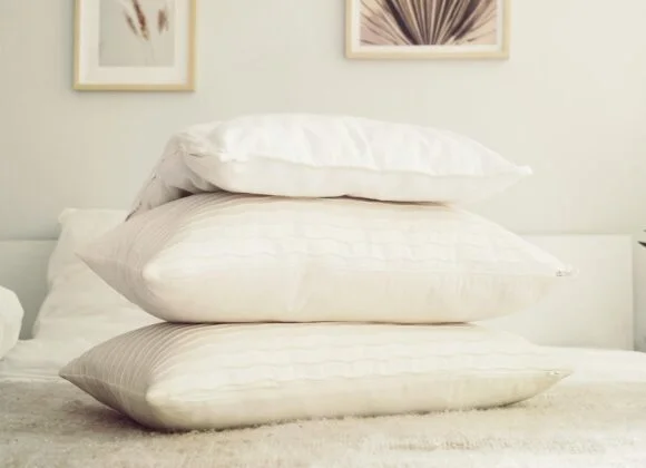 Three white pillows are stacked on a beige bed, with framed artwork hanging on a light-colored wall in the background.