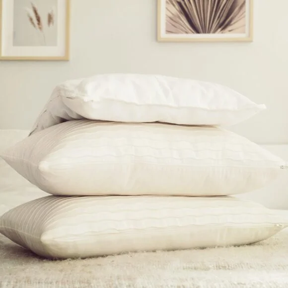 Three white pillows are stacked on a beige bed, with framed artwork hanging on a light-colored wall in the background.