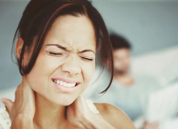 A woman grimaces in pain while holding her neck with both hands. In the background, a blurred figure is seen sitting, possibly reading a newspaper.