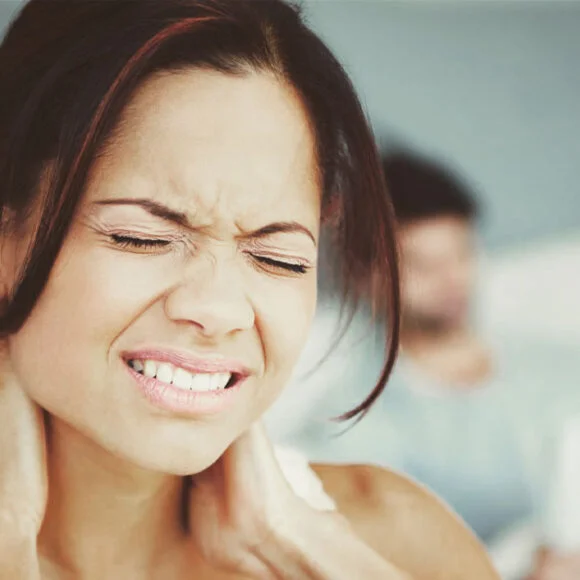 A woman grimaces in pain while holding her neck with both hands. In the background, a blurred figure is seen sitting, possibly reading a newspaper.